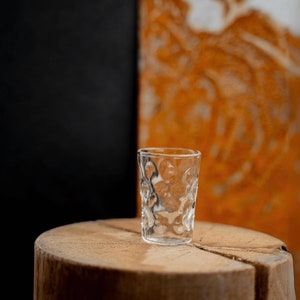 Peut inclure: Un verre &agrave; shot en verre transparent avec une surface textur&eacute;e pos&eacute; sur une surface en bois. Le verre est vide et a un fond arrondi.