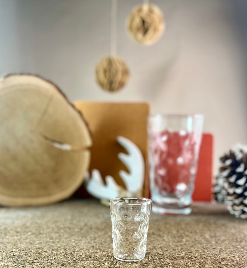 Peut inclure: Un verre &agrave; shot en verre transparent avec un motif textur&eacute; en forme de bulles. Le verre est pos&eacute; sur une surface brune.