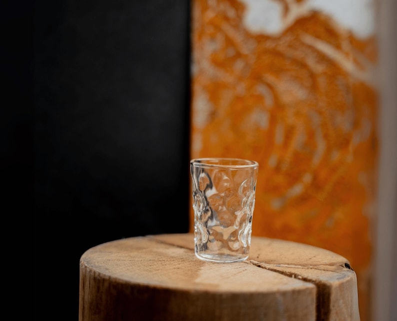 Peut inclure: Un verre &agrave; shot en verre transparent avec une surface textur&eacute;e pos&eacute; sur une surface en bois. Le verre est vide et a un fond arrondi.