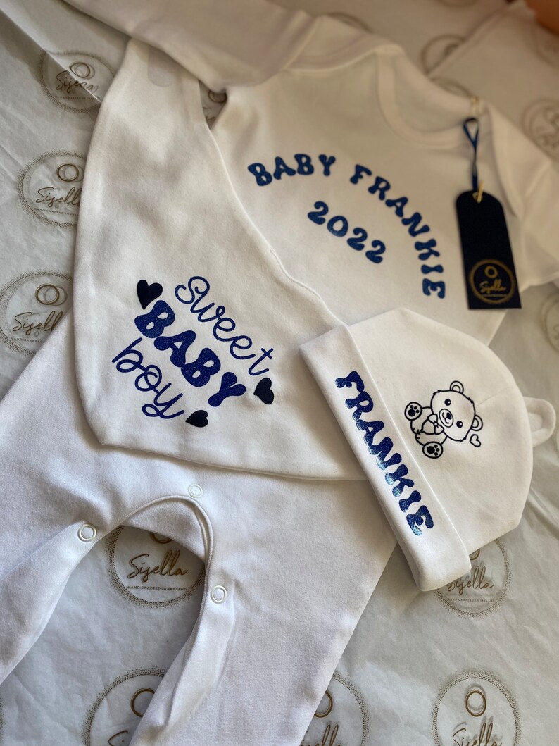 May include: A white baby onesie, bib and hat with blue text and a small bear graphic. The onesie says "Baby Frankie 2022". The bib says "Sweet Baby Boy" with hearts. The hat says "Frankie" with a bear graphic.