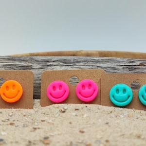 Könnte beinhalten: Eine Auswahl an Smiley-Ohrsteckern in leuchtenden Farben: Orange, Pink und Türkis. Jeder Ohrring ist auf einer kleinen, braunen Ohrringkarte befestigt. Die Ohrringe werden auf einer sandigen Oberfläche mit einem Holz-Hintergrund präsentiert.