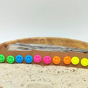 Könnte beinhalten: Eine Reihe bunter Smiley-Ohrstecker in Neongrün, Grün, Blau, Pink, Orange und Gelb. Die Ohrringe werden auf einer braunen Karte präsentiert, mit einer Holzschale und Treibholz im Hintergrund.