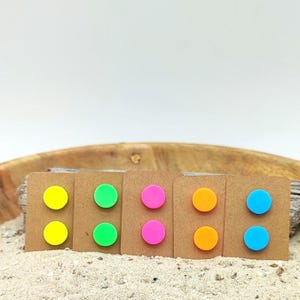 Könnte beinhalten: Eine Sammlung bunter, runder Ohrstecker, präsentiert auf kleinen braunen Pappkarten. Die Ohrringe sind paarweise in Gelb, Grün, Pink, Orange und Blau erhältlich. Der Hintergrund umfasst Sand und eine Holzschale.