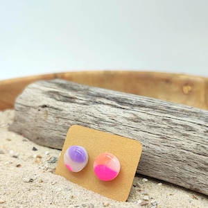 Könnte beinhalten: Zwei bunte Ohrstecker auf einer beigen Karte, vor dem Hintergrund von Sand und Treibholz. Ein Ohrring hat einen Farbverlauf aus Lila und Weiß, während der andere rosa und orangefarbene Farbtöne aufweist. Die Ohrringe sind rund und haben ein glänzendes Finish.