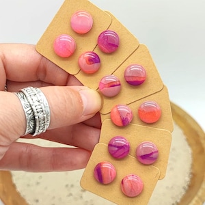 Könnte beinhalten: Fünf Paar runde Ohrstecker, die auf einzelnen braunen Pappquadraten präsentiert werden. Jeder Ohrstecker hat eine glatte, glänzende Oberfläche mit wirbelnden Mustern in Rosa-, Magenta-, Korallen- und Orangetönen, die einen lebendigen Effekt erzeugen.