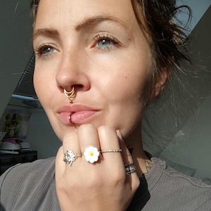Könnte beinhalten: Eine Person mit mehreren Ringen, darunter ein silberner Schmetterlingsring und ein weißer Gänseblümchenring mit gelber Mitte. Die Person hat auch einen Lippenring und mehrere Nasenpiercings mit Goldschmuck.