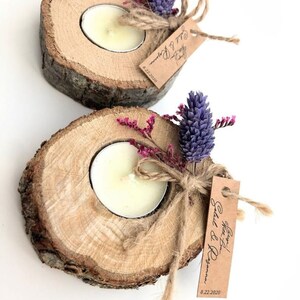 May include: Two rustic wooden candle holders with a tea light candle in each. The holders are decorated with dried lavender and pink flowers, and a small brown tag with the text "Save the Date!" and the date "8.22.2020".