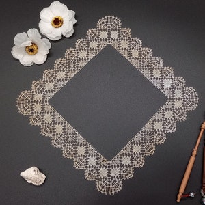 Op de afbeelding: Een vierkante, crèmekleurige kanten doily met een gedetailleerd, opengewerkt ontwerp. Twee witte papieren bloemen met gele centra en een kleine zeeschelp zijn rond de doily gerangschikt. Twee houten klosjes zijn ook aanwezig op een donkergrijze ondergrond.