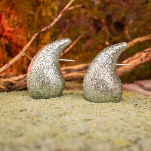 Può includere: Un paio di orecchini a bottone a forma di goccia argentati e glitterati. Gli orecchini hanno un perno per orecchie forate e sono posti su uno sfondo naturale con ramoscelli e muschio.