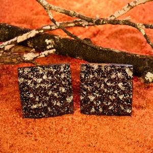 Puede incluir: Dos pendientes cuadrados con un diseño moteado en marrón oscuro y blanco. Se presentan sobre un fondo naranja texturizado, con una rama marrón oscuro en el fondo. Un accesorio único.