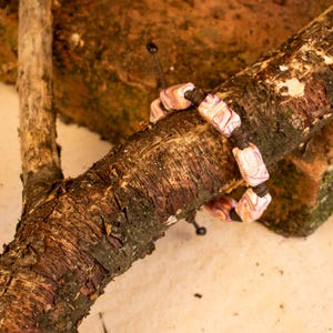 May include: A handmade bracelet with rectangular, marbled beads in shades of pink, white, and purple. The beads are strung on a dark brown cord. The bracelet is displayed on a textured, brown tree branch.
