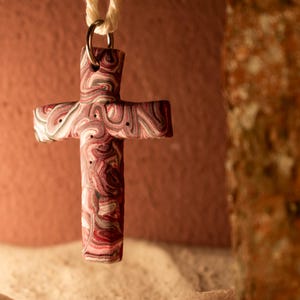 Può includere: Un ciondolo a forma di croce marmorizzata con vortici rossi, bianchi e neri. La croce è sospesa a un cordino color crema e a un anello color argento. Il ciondolo ha un aspetto strutturato su uno sfondo sfocato.