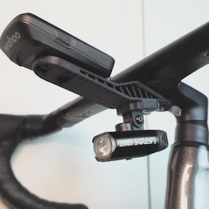 May include: A black 3D-printed bicycle mount attached to the handlebars, featuring a Wahoo Elemnt cycling computer on the top surface and a black Lezyne 500XL headlight mounted underneath the bracket, positioned on a road bike stem.