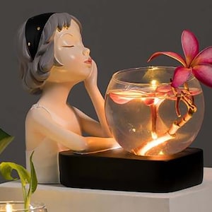 May include: A white porcelain figurine of a woman with a black headband, holding a glass bowl filled with water and a pink flower. The bowl is lit from within, creating a warm glow.