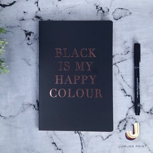 Może przedstawiać: Czarny notes z napisem "BLACK IS MY HAPPY COLOUR" w kolorze r&oacute;żowego złota. Notes leży na białym marmurowym blacie.