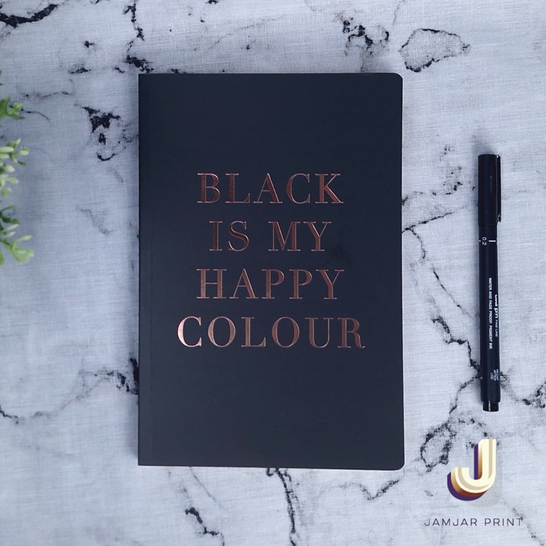 Może przedstawiać: Czarny notes z napisem "BLACK IS MY HAPPY COLOUR" w kolorze r&oacute;żowego złota. Notes leży na białym marmurowym blacie.