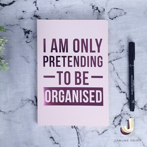 Może przedstawiać: R&oacute;żowy notes z tekstem "I am only pretending - to be - organised" w metalicznym fioletowym kolorze czcionki. Notes leży na marmurowej powierzchni z czarnym długopisem.