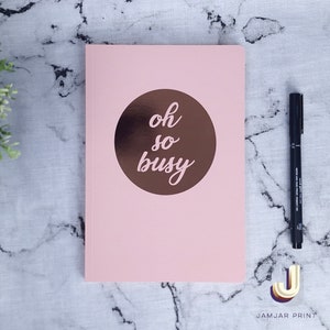 Może przedstawiać: R&oacute;żowy notes z miedzianym kołem na przedniej stronie. Koło zawiera napis "oh so busy" napisany kursywą.