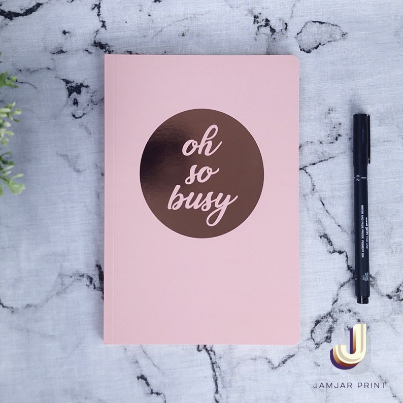 Może przedstawiać: R&oacute;żowy notes z miedzianym kołem na przedniej stronie. Koło zawiera napis "oh so busy" napisany kursywą.