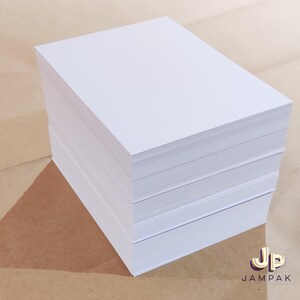 May include: A stack of bright white paper sheets, likely for printing or writing. The paper is neatly stacked, showing clean edges and a smooth surface. The background is a light brown color, with the logo "Jampak" in the bottom right corner.