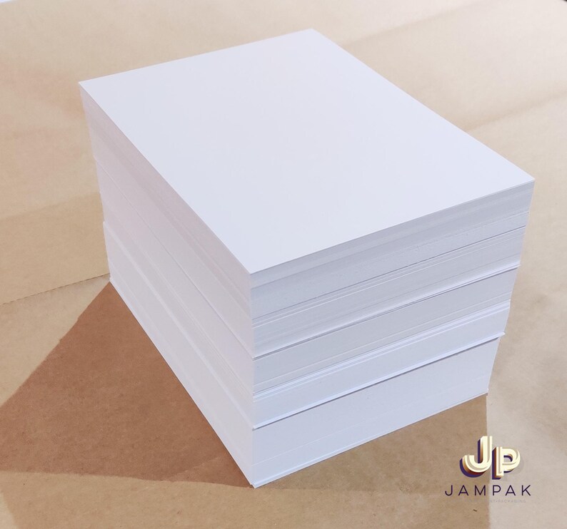 May include: A stack of bright white paper sheets, likely for printing or writing. The paper is neatly stacked, showing clean edges and a smooth surface. The background is a light brown color, with the logo "Jampak" in the bottom right corner.