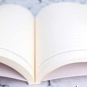 Może przedstawiać: Otwarty pusty notes z liniowanymi stronami, leżący na białym marmurowym blacie. Notes ma białą okładkę i biały grzbiet.