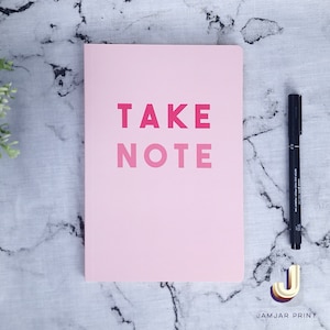 Może przedstawiać: R&oacute;żowy notes z napisem "TAKE NOTE" w kolorze r&oacute;żowym i czerwonym. Notes leży na marmurowej powierzchni obok czarnego długopisu.