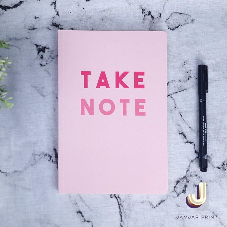 Może przedstawiać: R&oacute;żowy notes z napisem "TAKE NOTE" w kolorze r&oacute;żowym i czerwonym. Notes leży na marmurowej powierzchni obok czarnego długopisu.