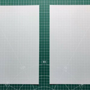 May include: Two sheets of tracing paper on a green cutting mat. The tracing paper is white and has a grid pattern. A black marker and a wooden stick are on the left side of the mat. A package of tracing paper is on the right side of the mat.