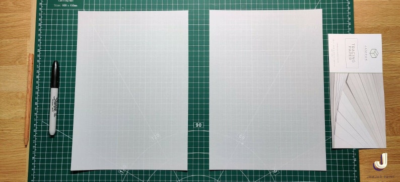 May include: Two sheets of tracing paper on a green cutting mat. The tracing paper is white and has a grid pattern. A black marker and a wooden stick are on the left side of the mat. A package of tracing paper is on the right side of the mat.