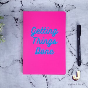 Może przedstawiać: Jasnor&oacute;żowy notes z napisem "Getting Things Done" w niebieskich literach. Notes leży na białym marmurowym blacie z czarnym długopisem.