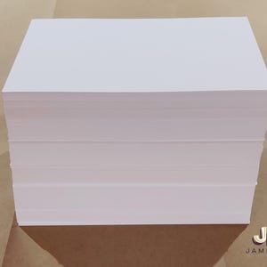 May include: A stack of white rectangular paper sheets, likely for printing or writing. The paper is neatly stacked, showing clean edges and a smooth surface. The image is taken from a slightly elevated angle, highlighting the stack's thickness and the crisp white color of the paper.