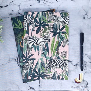 May include: Two notebooks featuring a pink and black zebra and tropical leaf design. The notebooks are stacked on top of each other. The top notebook is pink with black zebras and green leaves. The bottom notebook is black with green leaves and orange accents.