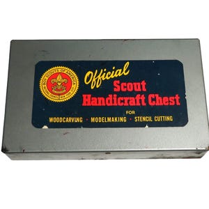 Puede incluir: Un cofre de artesanía oficial de los Boy Scouts, de metal gris rectangular. El cofre tiene una etiqueta azul con texto amarillo y rojo que dice "Official Scout Handicraft Chest" y el logotipo de los Boy Scouts of America. La etiqueta también enumera "Woodcarving, Modelmaking, Stencil Cutting."