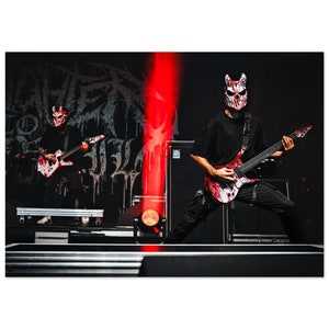 Könnte beinhalten: Zwei Musiker in roten und weißen Masken treten auf der Bühne auf. Der Musiker im Vordergrund spielt eine weiße E-Gitarre mit roten Farbschleudern. Der Musiker im Hintergrund spielt eine schwarze E-Gitarre mit roten Farbschleudern. Die Bühne ist mit roten und weißen Lichtern beleuchtet.