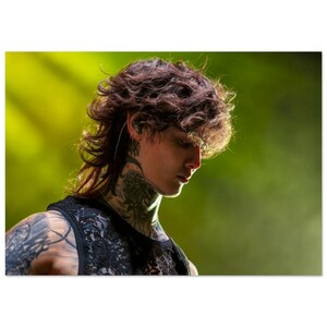 May include: A young man with long, curly brown hair and tattoos on his neck and arm. He is wearing a black sleeveless shirt and looking down.