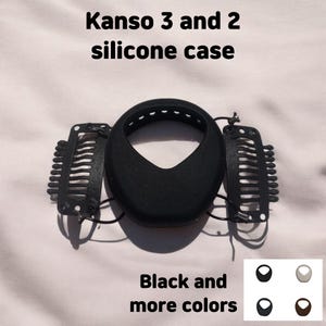 以下が含まれることがあります： Kanso 3 および 2 デバイス用の黒いシリコンケース。ケースは、中央の開口部と 2 つのクリップが付いたユニークな形状をしています。「Kanso 3 and 2 silicone case」と「Black and more colors」の文字が見えます。