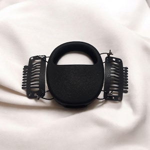 Puede incluir: Accesorio para el cabello negro con una pieza central ovalada y un asa. Dos clips metálicos con dientes en forma de peine están unidos a cada lado. Este accesorio está diseñado para asegurar el cabello, posiblemente para peinarlo o sujetar un peinado.