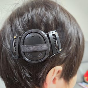 May include: A black circular electronic device secured to dark hair with two black metal hair clips. The device features a central oval housing with a textured horizontal strap across the middle, connected to the clips by thin black wires.