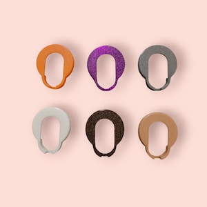 Puede incluir: Seis objetos ovalados en varios colores: naranja, morado brillante, gris, blanco, marrón oscuro y beige. Cada objeto tiene una forma redondeada con una abertura central, sobre un fondo rosa claro.