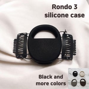 Puede incluir: Funda de silicona negra con el texto "Rondo 3 silicone case" y "Black and more colors". La funda tiene un diseño circular con dos clips negros adjuntos. Se muestran opciones de color adicionales en la esquina inferior derecha.