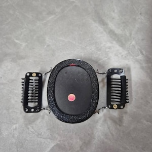 以下が含まれることがあります： 楕円形の中央部と2つのサイドクリップが付いた黒いヘアアクセサリー。中央部はテクスチャ加工された表面と小さな赤い点があります。クリップは黒色で、櫛のような歯が付いています。