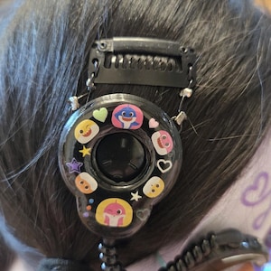 以下が含まれることがあります： カラフルな漫画のキャラクターと絵文字で飾られた、円形のデザインの黒いヘアアクセサリー。アクセサリーは、コームクリップとコイル状のコードで髪に固定されています。デザインには、サメ、ハート、星の画像が含まれています。