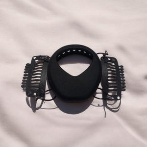 Puede incluir: Un moldeador de moño para el cabello negro con dos pinzas para el cabello negras. El moldeador de moño es un material redondo, negro y parecido a la espuma con agujeros alrededor del borde superior. Las pinzas para el cabello son de plástico con un diseño en forma de peine.