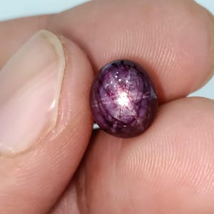 Może przedstawiać: Wypolerowany, owalny kamień szlachetny o głębokim fioletowym odcieniu i wzorze w kształcie gwiazdy. Klejnot trzymany jest między palcami, ukazując gładką powierzchnię i wewnętrzne odbicia światła. Kolor kamienia waha się od ciemnofioletowego do jaśniejszego, prawie różowego odcienia.
