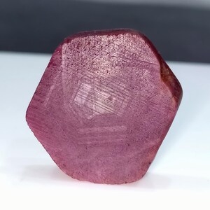 Peut inclure: Une pierre précieuse brute de rubis facettée, de couleur rose foncé. La pierre a une surface rugueuse et texturée et une forme géométrique. Le rubis est posé sur un fond blanc, avec un dégradé gris foncé et noir en arrière-plan.
