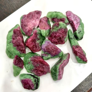 Puede incluir: Una colección de piedras en bruto sin pulir que presentan una combinación distintiva de inclusiones minerales en rojo magenta intenso y verde vibrante. Cada pieza tiene una forma irregular y dentada con una textura mate natural.