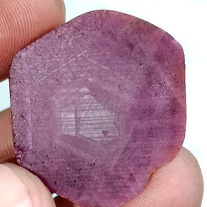 Puede incluir: Una gema de rubí hexagonal en bruto, con un tono magenta intenso. La piedra presenta un diseño grabado de una casa o edificio, agregando un elemento artístico único a la gema natural.