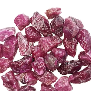 May include: A collection of rough-cut, deep red ruby gemstones. The raw rubies vary in size and shape, displaying a natural, unpolished texture. The gemstones are scattered against a white background, highlighting their rich color and crystalline structure.