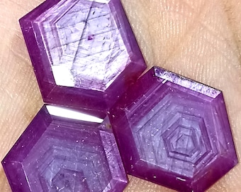 Puede incluir: Tres gemas hexagonales facetadas en tonos púrpura intenso. Cada gema tiene un diseño interior geométrico en capas. Las gemas se muestran sobre un fondo neutro, resaltando su color y cortes intrincados. Estas gemas podrían usarse para la fabricación de joyas.